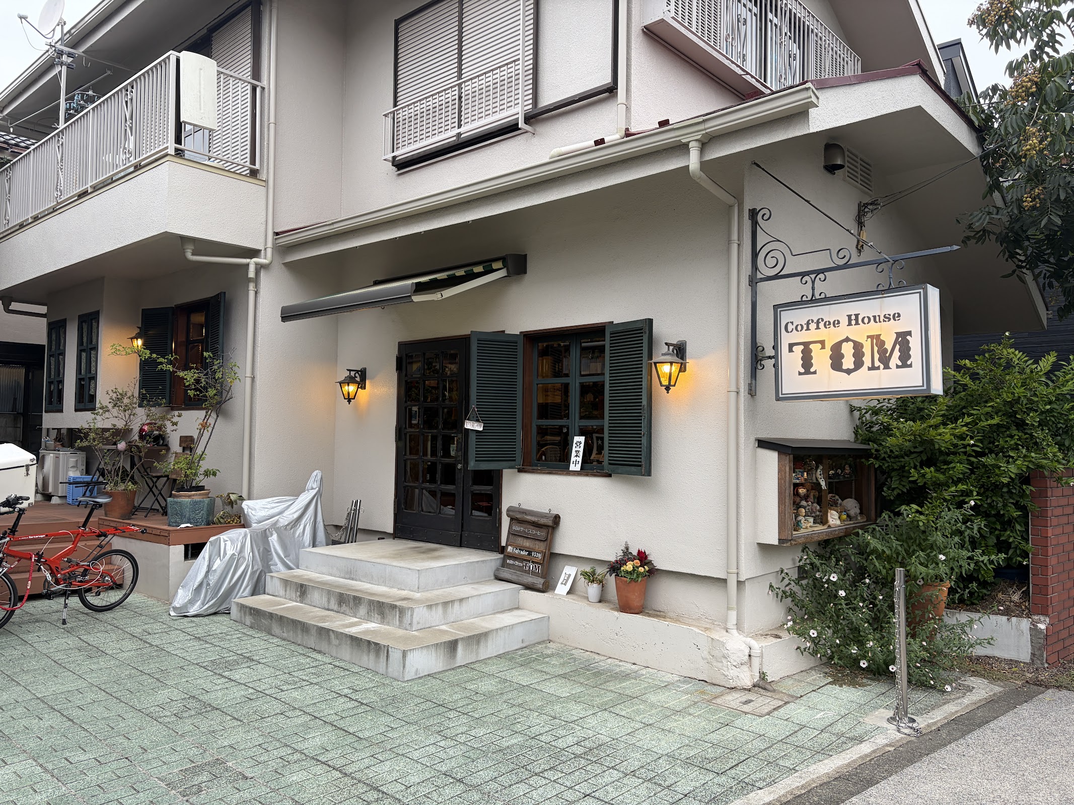 鉄製アイアンブラケットに吊るされたCoffee House TOM看板と一軒家外観、手前に本日のサービスコーヒー黒板